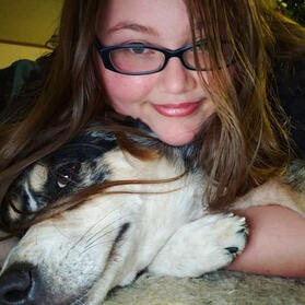 A picture of a caucasian person with brown hair, green eyes, and glasses hugging a white, grey, and black merle dog.